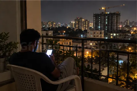 A man alone on an apartment balcony at night, lit by his phone, city lights behind