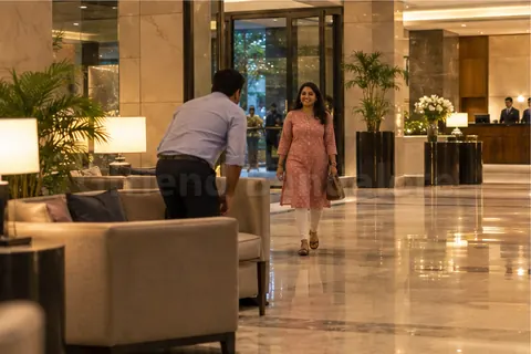 A woman in a pink kurta walking into a hotel lobby while a man gets up from the sofa to greet her