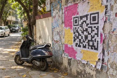 A torn pink and yellow poster with a big QR-code pattern on an old street wall, a scooter parked below